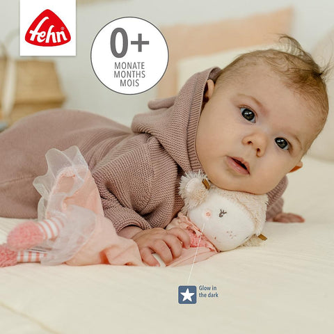 Ein Baby im Kapuzen-Outfit ruht auf einem Bett mit dem Schmusetuch Fee Plüschtier, das mit leuchtenden Elementen versehen ist und als Einschlafhilfe dient. In der Ecke sind das Etikett 0+ Monate und das Fehn-Logo zu sehen.