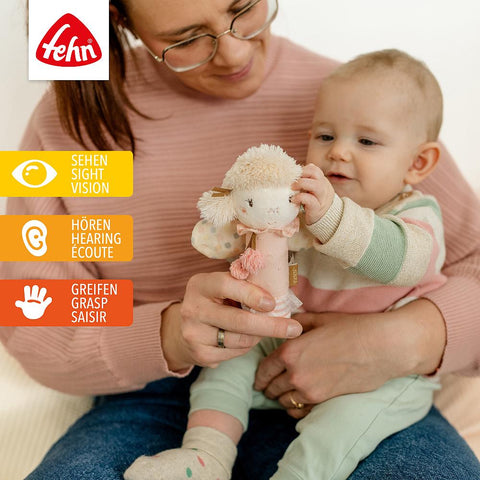 Ein Erwachsener hält ein Baby, das mit der Stabgreifling Fee spielt, einem weichen, im Dunkeln leuchtenden Spielzeug. Drei Symbole auf der linken Seite zeigen, dass es das Sehen, Hören und Greifen fördert. Das Fehn-Logo erscheint oben - ein perfektes Geschenk für die Kleinen.