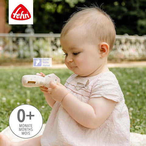 Ein Baby in einem hellrosa Kleid sitzt im Gras und hält die Mini-Rassel Maus. Das Bild zeigt das Fehn-Logo und hebt die Sinnesförderung für die Altersgruppe 0+ Monate/Monate/Mois hervor.
