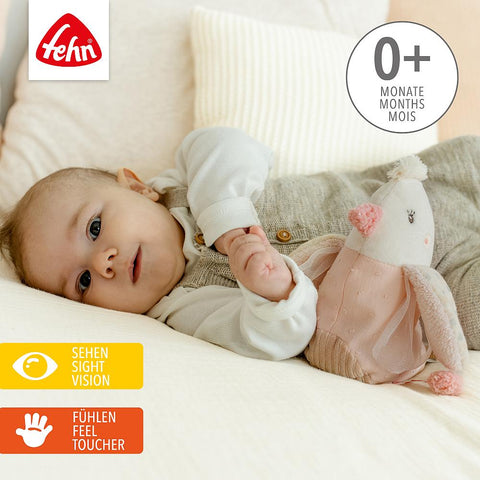 Ein Baby liegt auf einem Bett und hält das Wärmetier Vogel Plüschtier, geeignet für 0+ Monate. Das Spielzeug unterstützt die Seh- und Tastentwicklung und spendet mit seiner Traubenkernfüllung sanfte Wärme.