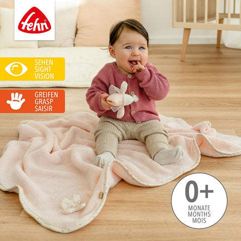 Ein Baby sitzt auf der weichen, rosafarbenen Kuscheldecke Vogel und hält ein Spielzeug in der Hand. Auf dem Bild sind "0+ Monate" und Symbole für Sehen und Greifen abgebildet.