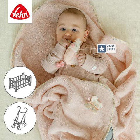 Ein Baby liegt auf einer weichen, rosafarbenen Kuscheldecke Vogel und hält ein im Dunkeln leuchtendes Plüschtier in der Hand, mit Abbildungen von Krippe und Kinderwagen, Vogelmotiv-Design und dem Fehn-Logo in der Ecke.