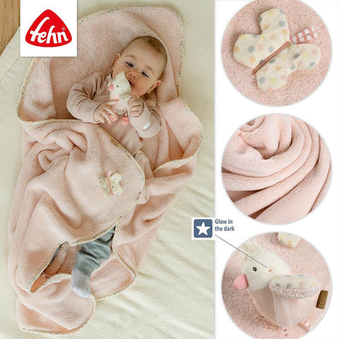 Ein Baby liegt auf einer rosa Kuscheldecke von Vogel und hält ein Stofftier. Die Einsätze heben die Decke, das Leuchten im Dunkeln, das Vogelmotiv und den Plüschschmetterling hervor und zeigen das Logo der Marke Fehn.