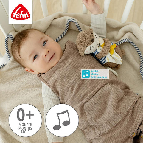 Ein Baby in einem Kinderbettchen trägt einen braunen Schlafsack und hält die Fehn Mini-Spieluhr Otter. Das Fehn-Logo und das beruhigende Spieluhr-Symbol zeigen, dass sie für das Alter von 0+ Monaten geeignet ist.