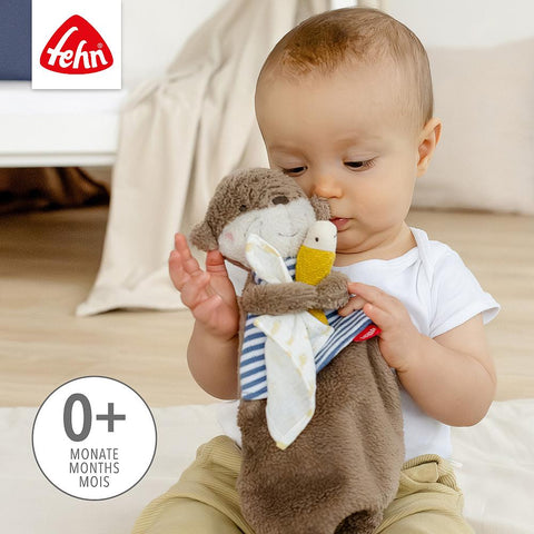 Ein Baby im weißen Hemd hält und erkundet das Schmusetuch Otter, ideal zum Kuscheln, Spielen und Trösten. Das Bild zeigt den Text "Fehn" und "0+ Monate Mois".