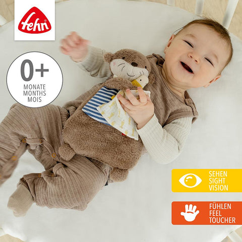 Ein lächelndes Baby liegt auf einer weißen Fläche und hält das Wärmetier Otter mit Kirschkernfüllung. Das Bild zeigt das Fehn-Logo und Etiketten für die Altersgruppen 0+, Sehen und Tasten.