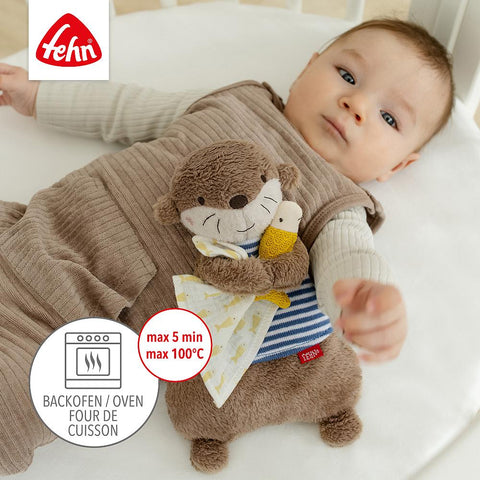 Ein Baby liegt in einem Kinderbett und hält das Wärmetier Otter mit Kirschkernfüllung von Fehn in der Hand, das einen niedlichen Otter und einen Vogel zeigt; das Fehn-Logo und die Heizanleitung für das Kirschkernkissen sind sichtbar.
