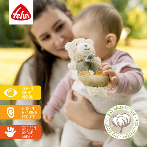 Eine Frau hält ein Baby, das mit dem Ring-Greifling Bär fehnNATUR kuschelt, einem motorischen Spielzeug aus Bio-Baumwolle. Piktogramme zeigen, dass es das Sehen, Hören und Greifen von Babys und Kleinkindern fördert. Das Bio-Baumwoll-Siegel ist ebenfalls sichtbar.