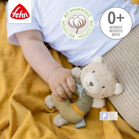 Baby mit dem Ring-Greifling Bär fehnNATUR, einem kleinen Plüschbären aus Bio-Baumwolle, liegend auf einer senfgelben Decke. Ideales Motorik-Spielzeug für Babys und Kleinkinder, geeignet für das Alter ab 0 Monaten.