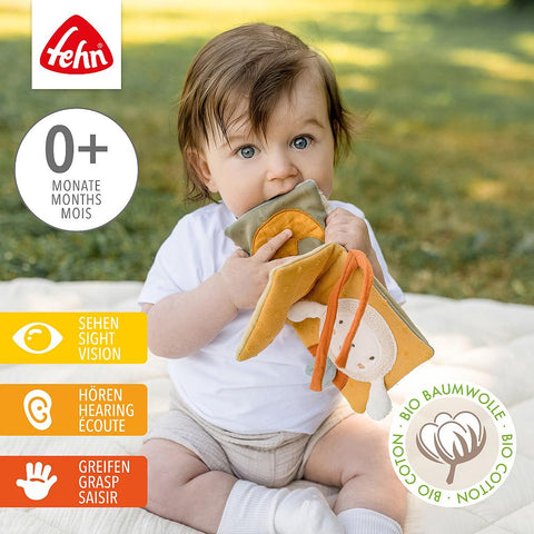 Ein Baby sitzt im Freien auf einer Decke und hält und kaut auf dem weichen Stoffbuch Schaf FehnNATUR. Piktogramme heben seine Vorteile hervor - es fördert das Sehen, Hören und Greifen - und ein Etikett zeigt, dass es aus Bio-Baumwolle hergestellt ist.