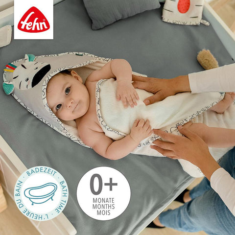 Ein Baby, eingewickelt in ein Kapuzenbadetuch Zebra, liegt auf einem Bett, während ein Erwachsener es sanft zudeckt. Das Fehn-Logo und das Etikett "Badezeit 0+ Monate" sind sichtbar.
