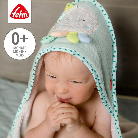 Ein Baby, das das Kapuzenbadetuch Oktopus mit einem Plüsch-Oktopus auf der Kapuze trägt, sitzt im Haus, die Hände vor dem Mund. Das Fehn-Logo und 0+ Monate sind sichtbar.