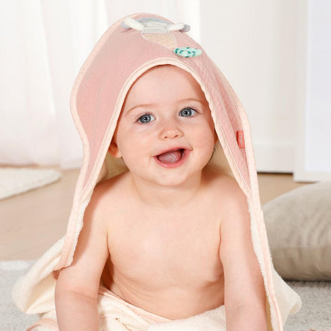 Ein lächelndes Baby mit einem Kapuzenbadetuch Meerjungfrau auf dem Kopf sitzt auf dem Boden und blickt mit offenem Mund in die Kamera.