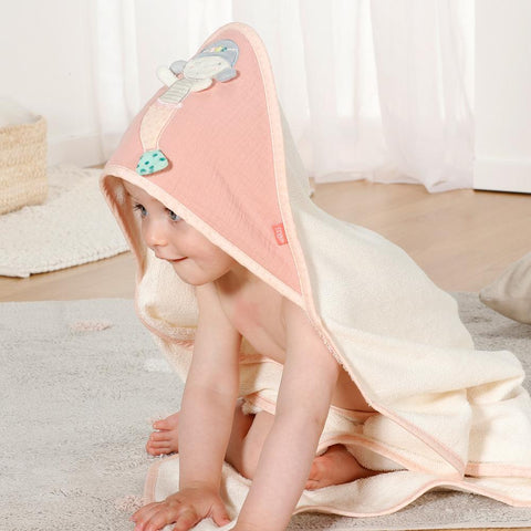 Ein Baby, eingewickelt in ein creme- und rosafarbenes Kapuzenbadetuch Meerjungfrau mit Tiergesicht, sitzt auf einem Teppich in einem lichtdurchfluteten Raum.