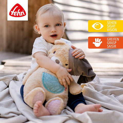 Ein Baby sitzt auf einer Decke im Freien und umarmt seinen XL Kuschelbär als kuscheligen Kuschelfreund. In der rechten oberen Ecke erscheinen das Fehn-Logo und die Symbole für Sehen und Greifen.