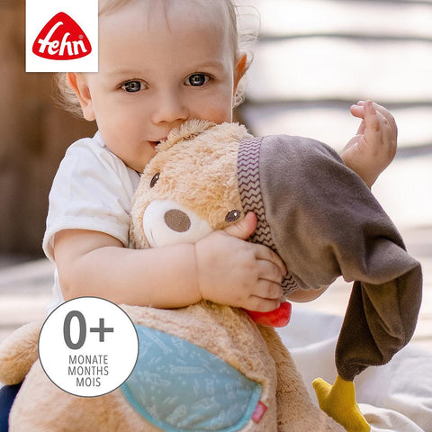Ein Baby kuschelt sich an einen XL-Kuschelbär mit Mütze; das Bild zeigt das Fehn-Logo und ein Etikett mit der Aufschrift 0+ Monate Mois.