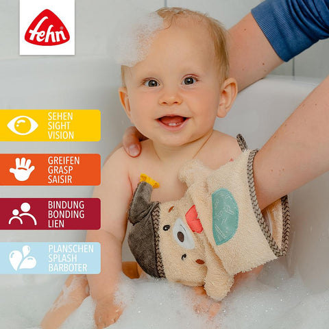 Ein lächelndes Baby sitzt in einer seifigen Badewanne, gehalten von einem Erwachsenen, der den Waschhandschuh Bär benutzt. Icons heben die Funktionen Sehen, Greifen, Binden und Spritzen auf Deutsch und Englisch hervor.