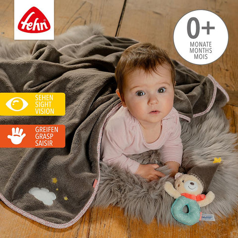 Ein Baby liegt auf einem Holzboden unter der Kuscheldecke Bär und hält einen Plüschbären. Die Kuscheldecke ist für Kinder ab 0 Jahren geeignet und fördert die Entwicklung des Sehens und Greifens.