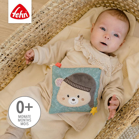 Ein Baby in Beige liegt in einem geflochtenen Korb und hält das Kirschkernkissen Bär. Der Text auf dem Bild besagt, dass es für Babys und Kleinkinder ab 0 Monaten geeignet ist.
