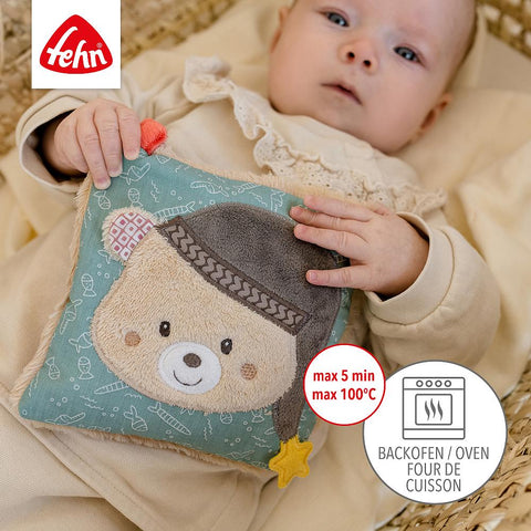 Ein Baby in einem Weidenkorb hält das Fehn Kirschkernkissen Bär - ein quadratisches Kirschkernkissen mit einem Bärengesicht, ideal als Wärmebegleiter für Babys und Kleinkinder. Unten rechts steht die Anleitung zum Erwärmen im Ofen, oben links das Fehn-Logo.