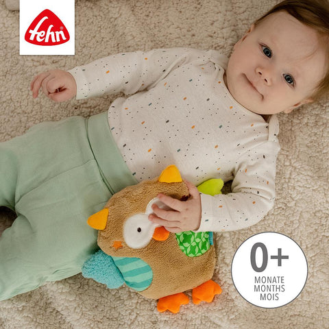 Ein Baby liegt auf einer beigen Fläche, bekleidet mit einer hellgrünen Hose und einem weißen Hemd, und kuschelt sich an das Wärmekissen Eule aus Plüsch. Das Fehn-Logo und der Text "0+ Monate" sind auf dem Bild zu sehen.