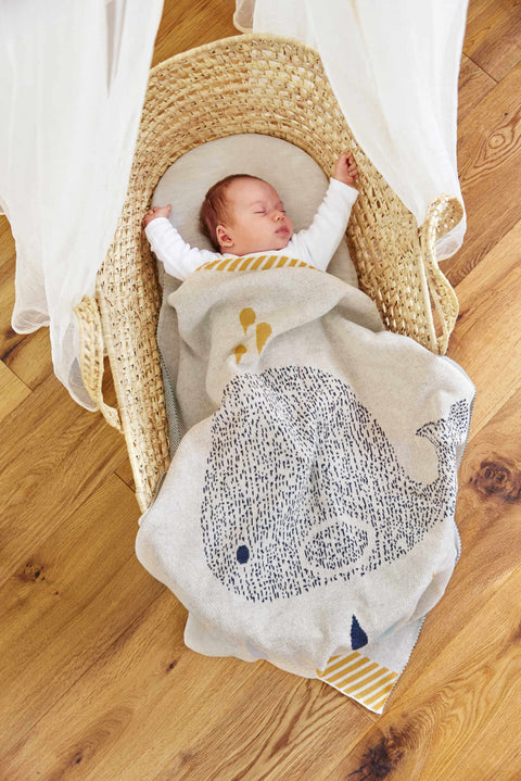Ein Baby schläft in einem gewebten Stubenwagen, eingehüllt in die Strickdecke - Wal (75x100cm) mit blauem Walmotiv. Der Stubenwagen ruht auf dem Holzboden eines gemütlichen Kinderzimmers.