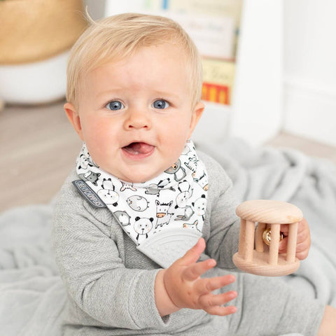 Ein blondes Baby in einem grauen Outfit und einem Neckerchew Zahnen Dribble Lätzchen - Panda Pals mit Tiermotiven sitzt auf einer Decke, hält einen hölzernen Beißring und schaut in die Kamera.