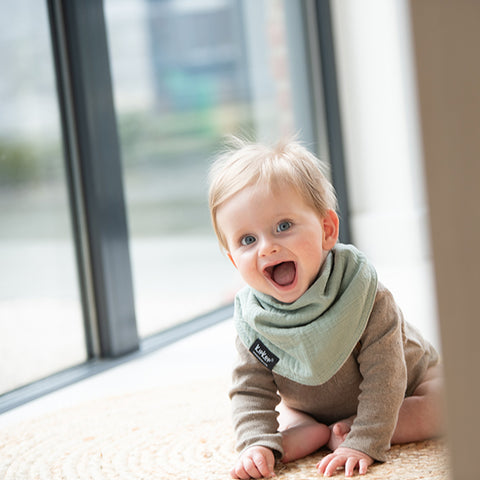 Ein lächelndes Baby mit hellem Haar sitzt auf einem gewebten Teppich vor einem großen Fenster, trägt ein beiges Outfit und das 3 in 1 Lätzchen (Spucktuch, Latz, Halstuch) in hellgrün.