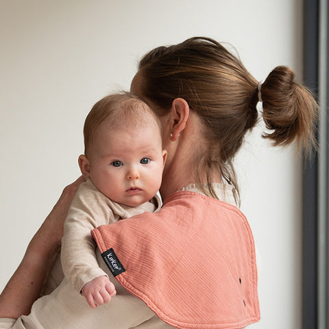 Eine Frau hält ein Baby über der Schulter, drapiert mit dem lachsfarbenen 3 in 1 Lätzchen (Spucktuch, Latz, Halstuch) - Sienna. Das kuschelige Baby schaut zufrieden in die Kamera.