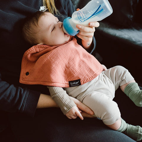 Ein Erwachsener füttert ein Baby mit einem Fläschchen, während das leicht bekleidete Baby ein sienafarbenes KipKep 3-in-1-Lätzchen (Spucktuch, Latz, Halstuch) trägt und bequem auf dem Schoß des Erwachsenen liegt.