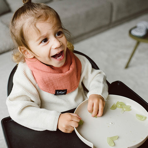 Ein kleines Kind mit einem Zopf lächelt an einem Tisch, es trägt einen weißen Pullover und das 3 in 1 Lätzchen (Spucktuch, Latz, Halstuch) - Sienna, mit aufgeschnittenen grünen Trauben auf einem Teller.