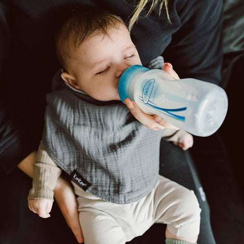 Ein Baby, das ein KipKep 3 in 1 Lätzchen (Spucktuch, Latz, Halstuch) in Grau trägt, trinkt Milch aus einer Flasche, während es von einem schwarz gekleideten Erwachsenen gehalten wird.