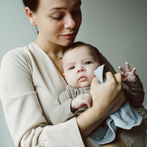 Eine Frau hält ein Baby im Haus und stützt das Kind sanft mit dem 3 in 1 Lätzchen (Spucktuch, Latz, Halstuch) in grau. Das Baby schaut in Richtung der Kamera.