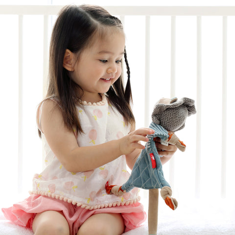 Ein kleines Mädchen in weißem und rosafarbenem Outfit sitzt im Haus und hält lächelnd das Schnuffeltuch - Lulo Lumpo, eine löwenförmige Kuscheldecke, die perfekt für Neugeborene und Kleinkinder ist.