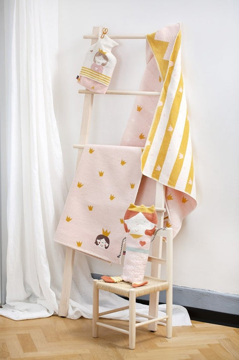 Auf einer Holzleiter liegen rosa und gelbe Handtücher mit Kronen- und Prinzessinnenmotiven. Eine passende LENA BW Decke Prinzessin und ein Handtuch liegen auf einem kleinen geflochtenen Stuhl auf einem Holzboden neben weißen Vorhängen.