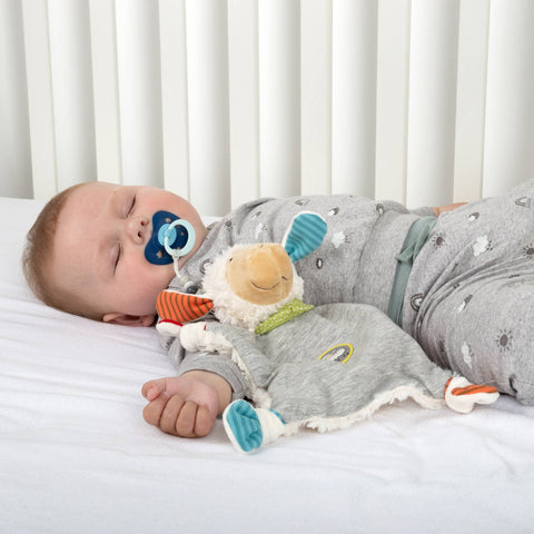 Baby schlafend auf einem weißen Laken mit Schnuller, in einem grauen Strampler, mit dem Schnuffeltuch Schaf, Boller Schäfle als Babyspielzeug, und vor weißen Lattenrosten liegend - Förderung der emotionalen Entwicklung.