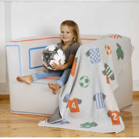 Ein kleines Mädchen sitzt auf einer weißen Couch und hält einen Fußball in der Hand. In der Nähe befindet sich das JUWEL Kinderdecke Fußball Trikot (100 x 140 cm) - perfekt für kleine Fußballfans.