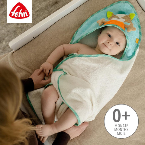 Baby eingewickelt in ein saugstarkes Frottee Kapuzenbadetuch Fuchs, liegend auf einer beigen Fläche, während ein Erwachsener die Seite des Babys hält. Das Fehn-Logo und der Text 0+ Monate sind sichtbar.
