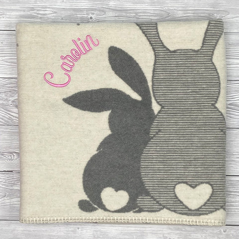 Die Jacquard Decke s.Oliver Junior - Häschen (75x100cm) aus Baumwollmischung zeigt zwei graue Bunny-Silhouetten, weiße Herzchen und ist personalisiert mit dem Namen Carolin in pinker Schrift auf hellem Holz.