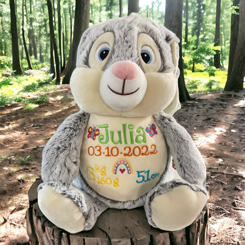 Clovis Brampton III, der Cubbies Hase, ein graues personalisiertes Plüschtier, sitzt im Wald auf einem Baumstumpf. Julias gestickte Geburtsdaten-Datum, Gewicht und Größe-stehen auf dem Bauch: ein liebevolles Geschenk.