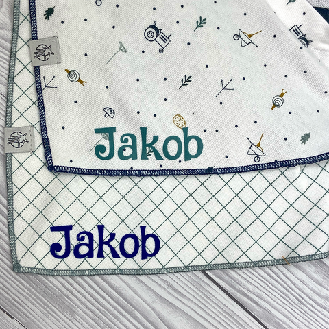 Auf einer Holzfläche liegen zwei Dreieckstücher Baby" aus Bio-Baumwolle - eines mit einem geometrischen Muster, das andere mit einem Garden Explorer Traktor-Motiv; beide sind mit dem Namen Jakob bestickt.