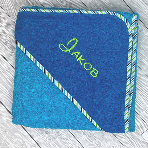 Ein Kapuzenbadetuch Aqua/Petrol 100x100cm aus Frottee mit gestreiftem Rand, personalisiert mit dem Namen Jakob in Grün auf der Kapuze, liegt auf einer hellen Holzoberfläche.