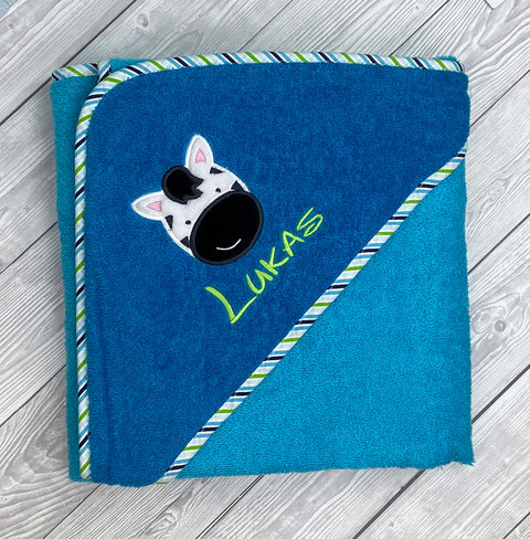 Das Kapuzenbadetuch Aqua/Petrol 100x100cm aus Frottee mit Zebra-Patch, Lukas-Stickerei und gestreiftem Rand ist stilvoll auf hellem Holz arrangiert.