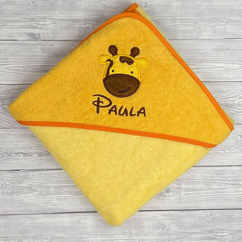 Kapuzenbadetuch Hellgelb/Gold 100x100cm aus Baumwolle für Kinder mit gesticktem Giraffenmotiv und dem Namen Paula auf der Kapuze, liebevoll auf heller Holzoberfläche arrangiert.