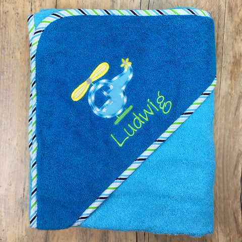 Ein gefaltetes Kapuzenbadetuch Aqua/Petrol 100x100cm mit gestreifter Bordüre, einem gestickten Hubschrauber und dem Namen Ludwig personalisiert auf der Ecke liegt auf einer Holzfläche.