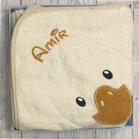 Das Sterntaler KBT - Edda Ente 100cmx100cm, ein cremefarbenes Baumwoll-Kapuzenbadetuch mit gesticktem Entengesicht und "Amir" in Gelb, präsentiert sich als niedliches Baby-Accessoire in einer Box auf einer Holzunterlage.