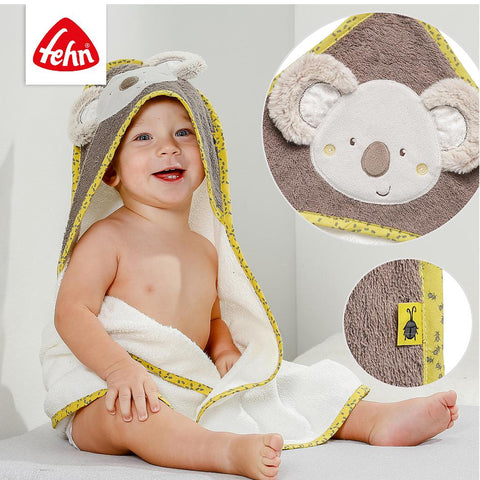 Ein lächelndes Baby wird in das Kapuzenbadetuch Koala eingewickelt. Es besteht aus weichem Frottee, einem niedlichen Koala-Gesicht mit grauen Ohren und einer detaillierten Applikation. Einsätze heben die Textur des Stoffes und das kleine Produktetikett hervor.