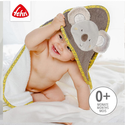 Ein lächelndes Baby ist in das Kapuzenbadetuch Koala mit einer niedlichen Koala-Applikation eingewickelt, neben dem Text 0+ Monate Mois und dem Fehn-Logo oben links.