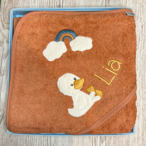Das orangefarbene Kapuzenbadetuch Edda Ente mit Motiven (100x100cm) ist aus 100% Baumwolle und hat eine Enten- und Regenbogenstickerei mit "Lia" in Gelb.