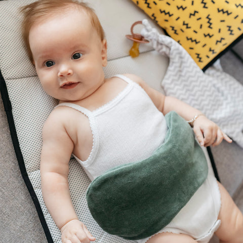 Ein Baby in einem weißen Strampler liegt auf einer Matte mit dem KipKep Wärmekissen - Calming Green aus Bio-Baumwolle auf dem Bauch für Koliken Linderung; Decken und Schnuller liegen daneben.